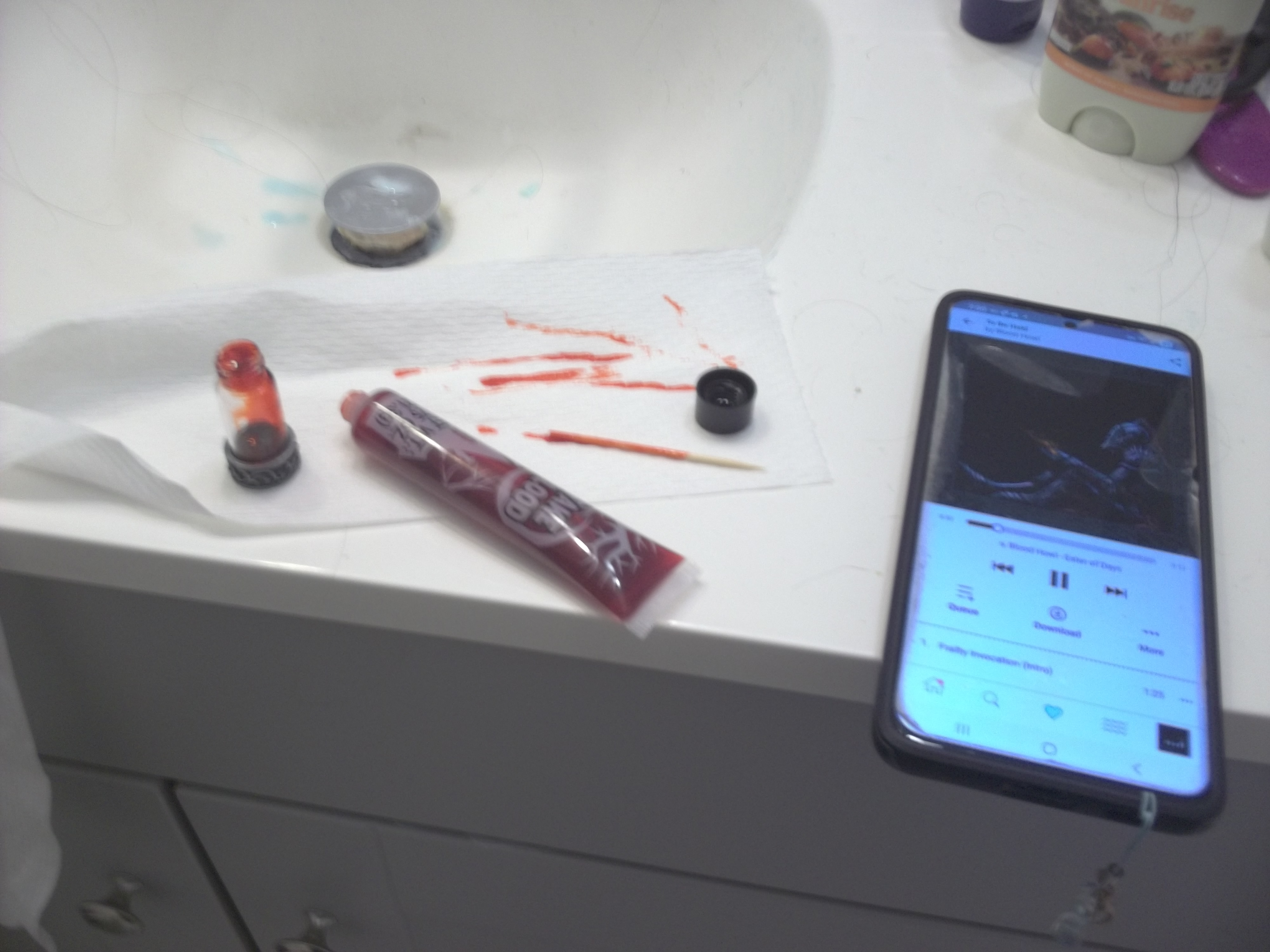 <image containing a mess on a bathroom counter with a vile of fake blood with the Bandcamp app open on a phone playing the song Eater of Days by Blood Howl>