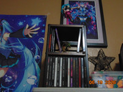 an upward view of a cd rack. the bottom storage compartment of the rack is full of cds, while the top only has a few and a mega rayquaza figure. on top of the rack is a hatsune miku figure, with two miku posters visible in the background