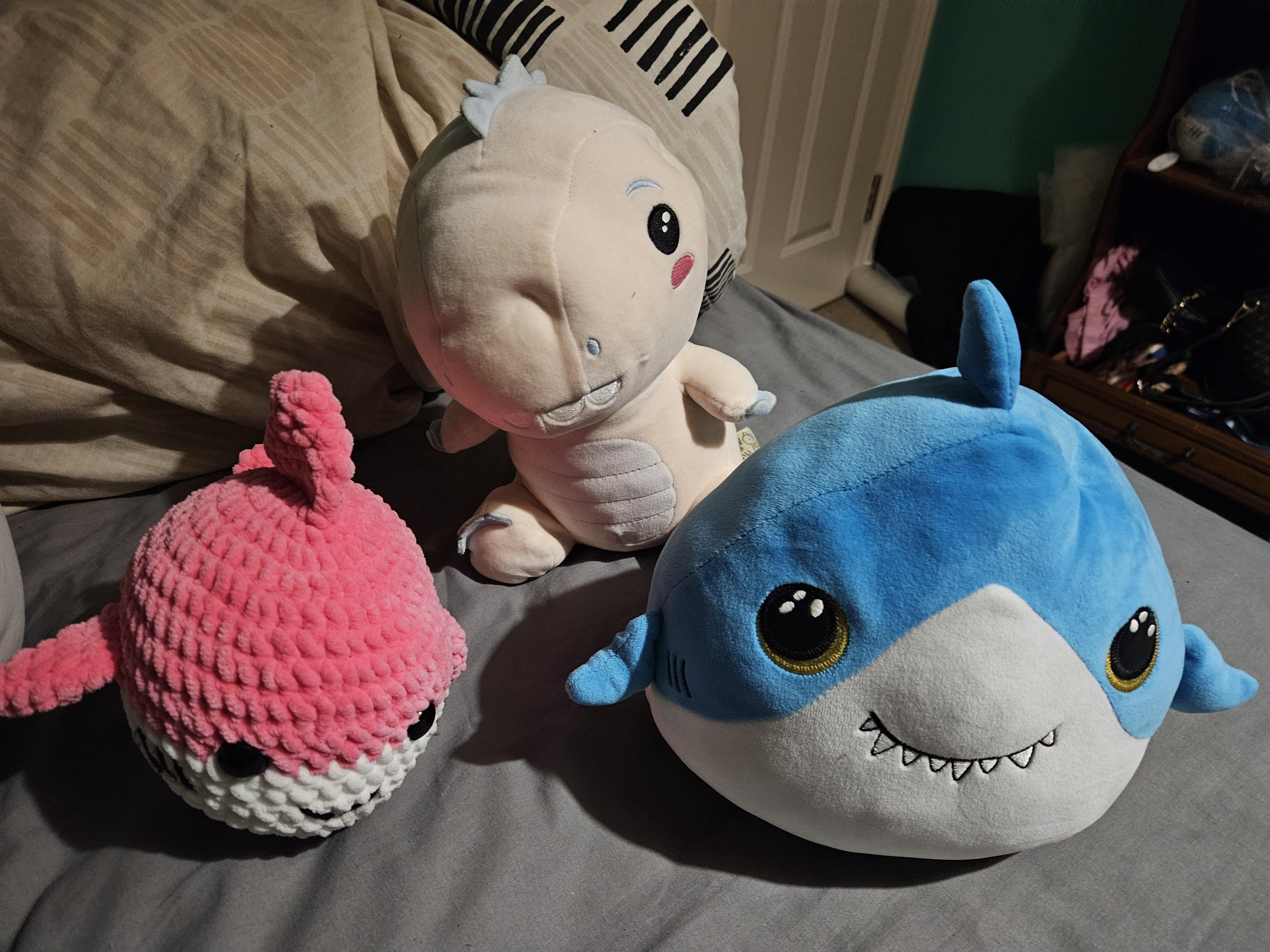 Left to right:a small round ball shaped crocheted pink shark, a bigger of the previously mentioned pink anthro dinoasuar, and a oval shaped silly shark with pointy teeth